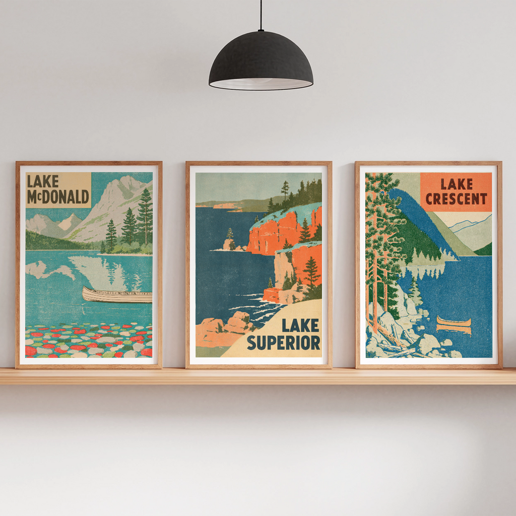 Three framed vintage-style travel posters showing Lake McDonald, Lake Superior, and Lake Crescent, displayed on a wooden shelf in a modern room setting with a floor lamp, hanging pendant light, and small potted plants.