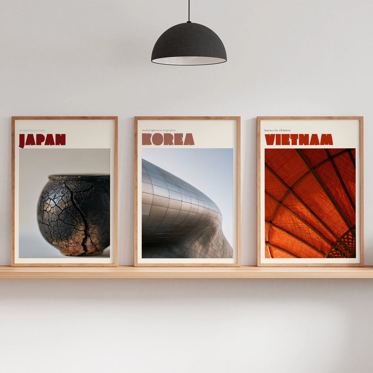 Three framed travel posters of Japan, Korea, and Vietnam displayed on a light wood floating shelf against a white wall, styled with a tripod floor lamp, a black pendant light, and small potted plants.