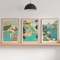 Three framed travel-style art prints depicting Lake Bled, Lac Danneycy, and Lake Bohinj are displayed on a wooden shelf in a modern room setting with a floor lamp, hanging pendant light, and small potted plants.