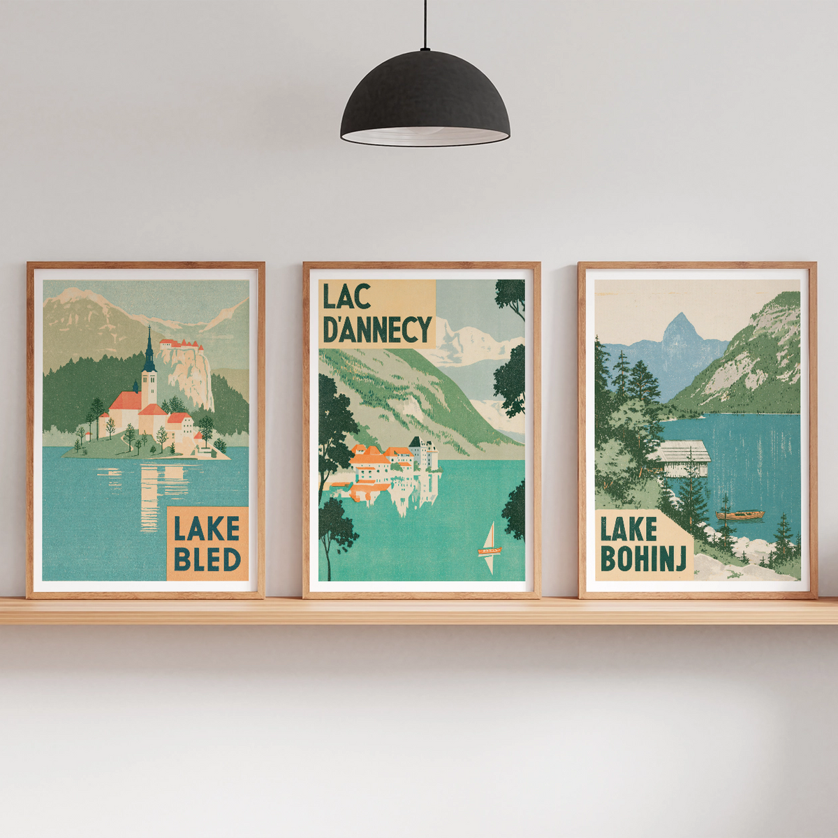Three framed travel-style art prints depicting Lake Bled, Lac Danneycy, and Lake Bohinj are displayed on a wooden shelf in a modern room setting with a floor lamp, hanging pendant light, and small potted plants.
