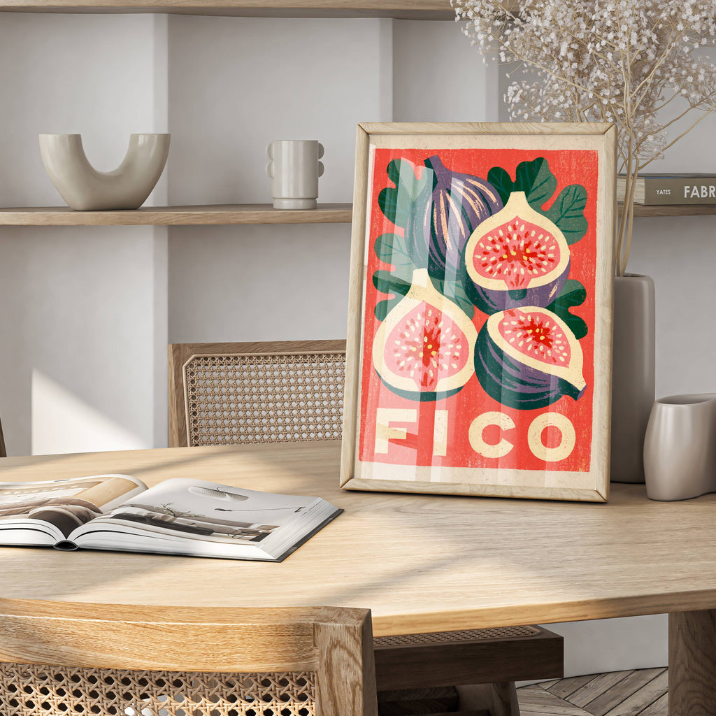 A framed vintage-style fig print with bold red, green, and cream colors stands on a light wood dining table in a modern interior setting with woven chairs, shelves, and an open magazine.