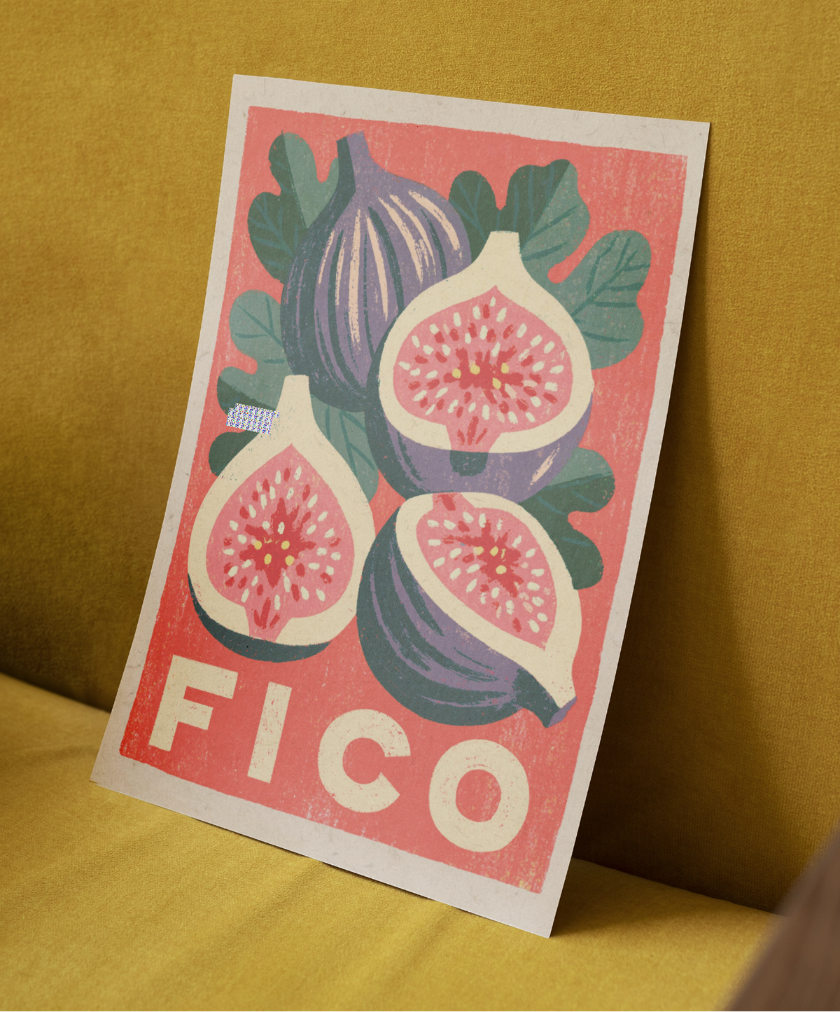 A vintage-style illustrated print featuring figs and the word “FICO” in bold cream letters on a pink background, leaning against a mustard yellow fabric surface.