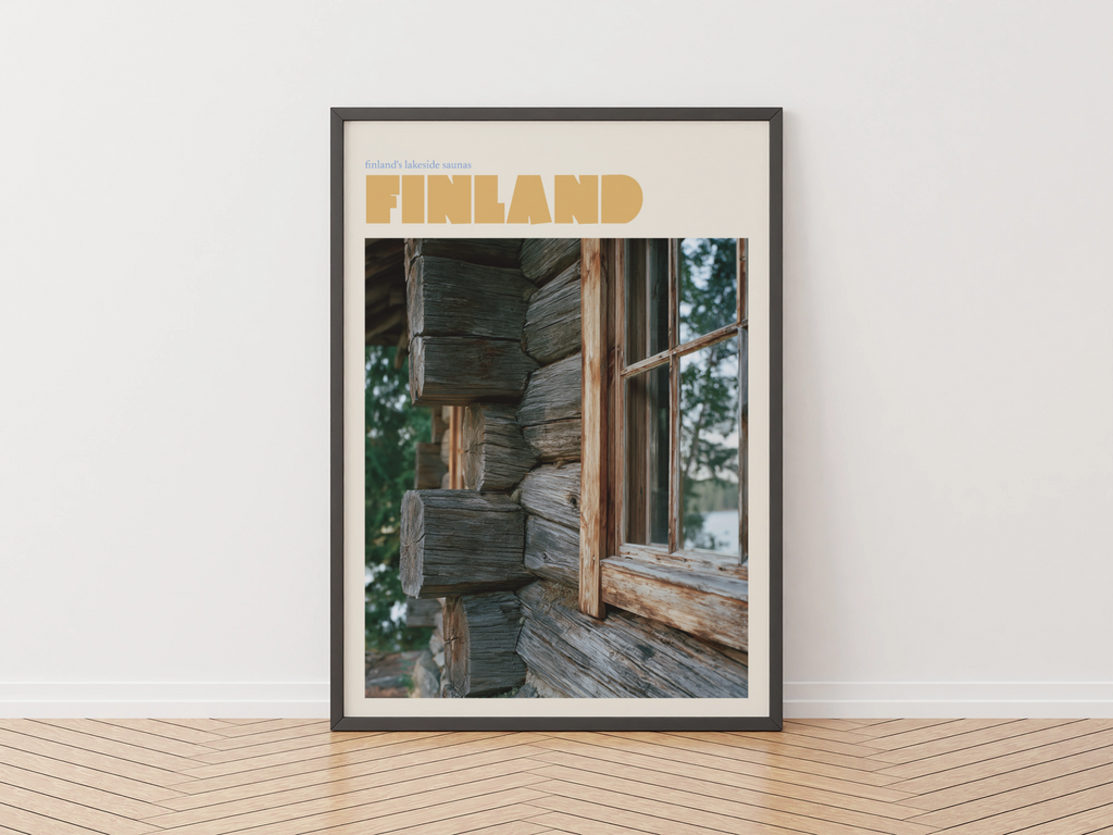 A framed travel poster of Finland leaning against a white wall on a light wood herringbone floor. The poster features a rustic log cabin window with the word 'FINLAND' in bold tan letters at the top.