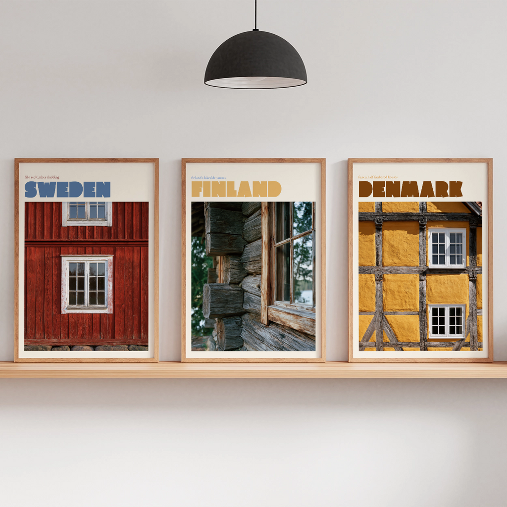 Three framed travel-style posters showing Sweden, Finland, and Denmark with rustic building imagery, displayed on a light wood shelf against a white wall in a styled interior with a floor lamp, hanging pendant, and small potted plants.