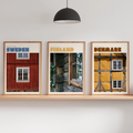 Three framed travel-style posters showing Sweden, Finland, and Denmark with rustic building imagery, displayed on a light wood shelf against a white wall in a styled interior with a floor lamp, hanging pendant, and small potted plants.