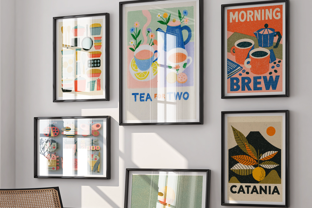 A styled interior room with six framed colorful art prints displayed on a white wall above a black chair. The prints feature retro-style illustrations including a tea set, morning brew coffee cups, a city skyline labeled Dallas, and a mountain labeled Catania.