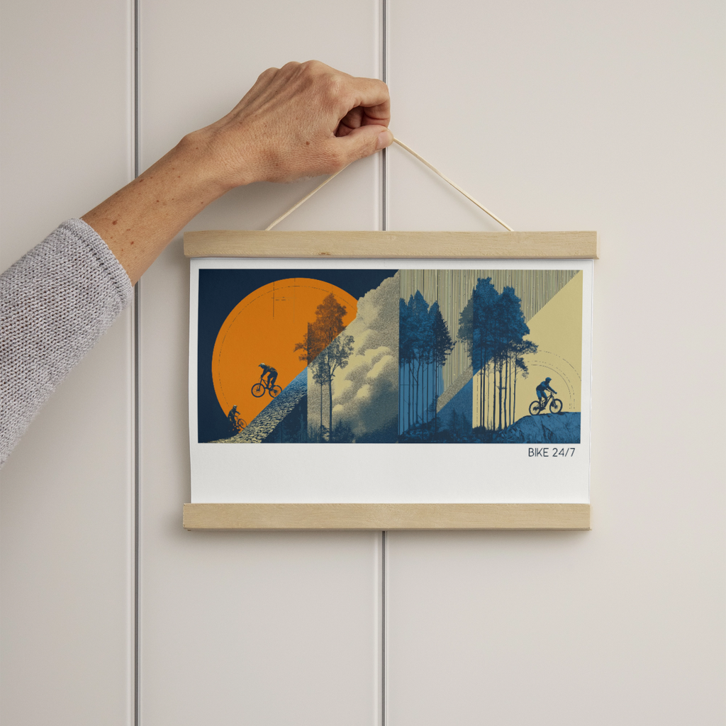 A hand holds a square framed art print hanging on a light gray paneled wall. The print features a stylized illustration of two mountain bikers against a large orange sun, blue trees, and clouds, with the text 'BKE 247' in the lower right corner.